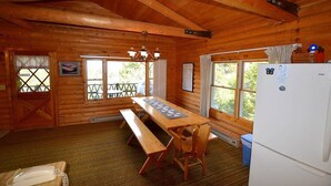 Eine Holzhütte mit Esstisch, Bänken, einem Kühlschrank und Fenstern mit Vorhängen.