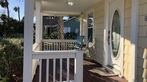 Une véranda couverte avec une balustrade blanche, une porte dotée d’une vitre et, au fond, une maison verte.