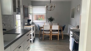 Eine moderne Küche mit weißen Schränken, einem Esstisch mit Stühlen, einer Pendelleuchte und einem Blick ins Freie durch ein Fenster.