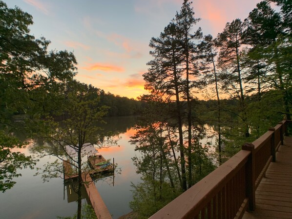 A wooden deck overlooking a calm lake with a dock and a boat, surrounded by tall trees and a sunset sky. - Water's Edge – Steps from the lake w/firepit, large entertaining spaces & more (Gainesville)