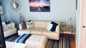 A living room with a beige sectional sofa, a small side table, a wall-mounted picture of a beach sunset, and a ceiling fan. - Milepost 9 KDH Condo Oceanside (Kill Devil Hills)
