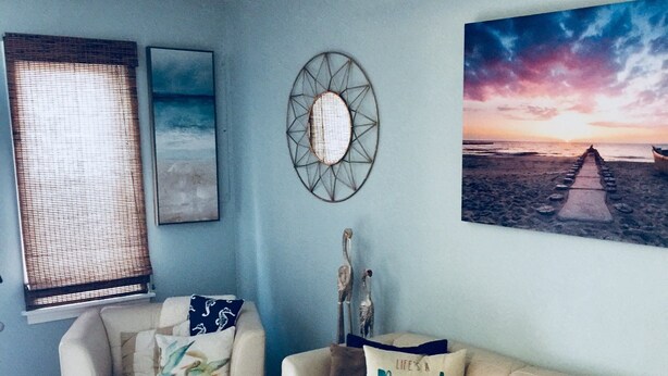 A living room with a beige sectional sofa, a white armchair, and a blue patterned rug. There is a ceiling fan, a wall-mounted mirror, and a painting of a beach scene. - Milepost 9 KDH Condo Oceanside (Kill Devil Hills)