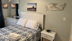 A bedroom with a bed, a nightstand, a dresser, a mirror, and wall art. - Milepost 9 KDH Condo Oceanside (Kill Devil Hills)