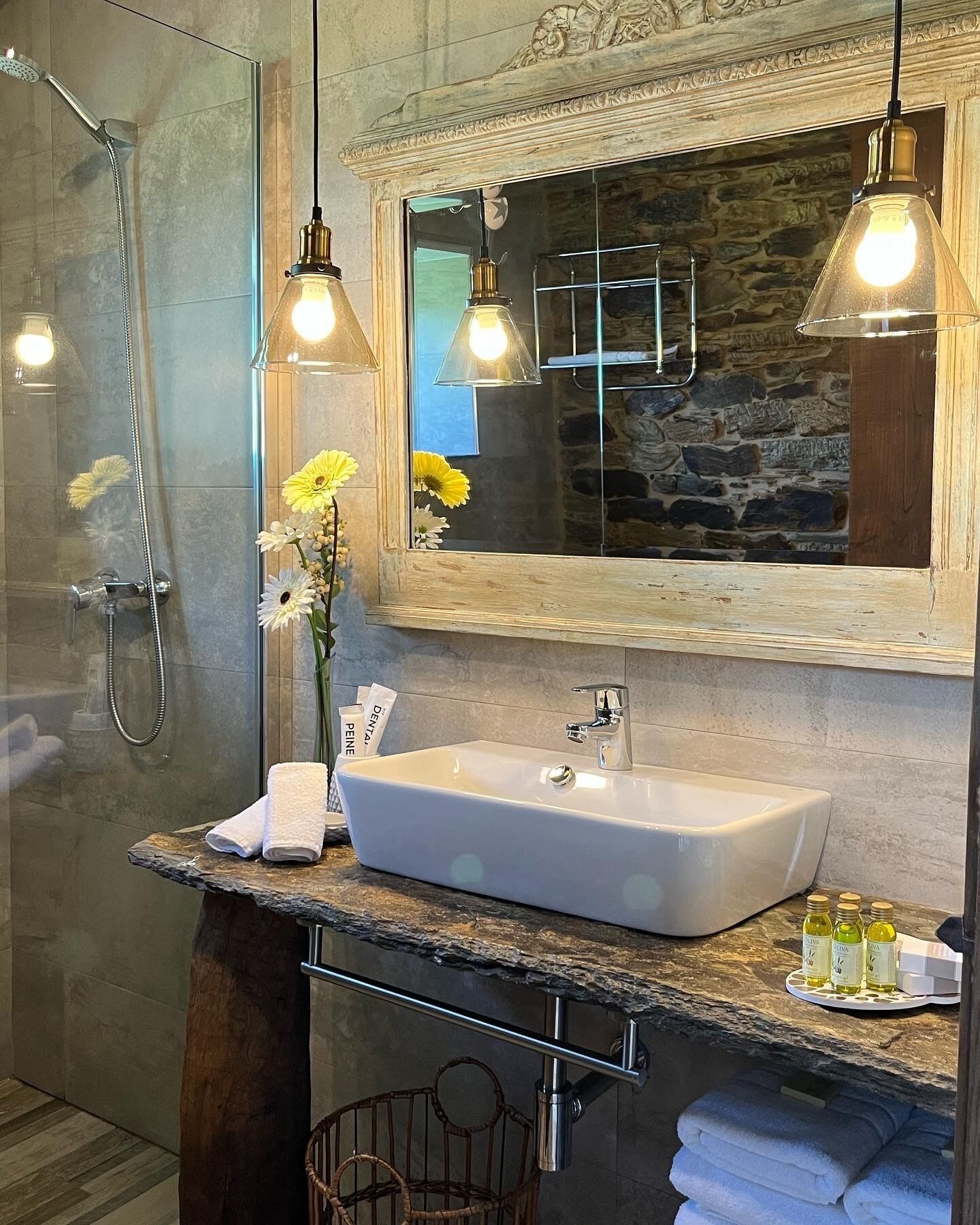 A modern bathroom with a stone countertop, a white sink, and a shower. There are two pendant lights, a mirror with a stone frame, and a vase with flowers.