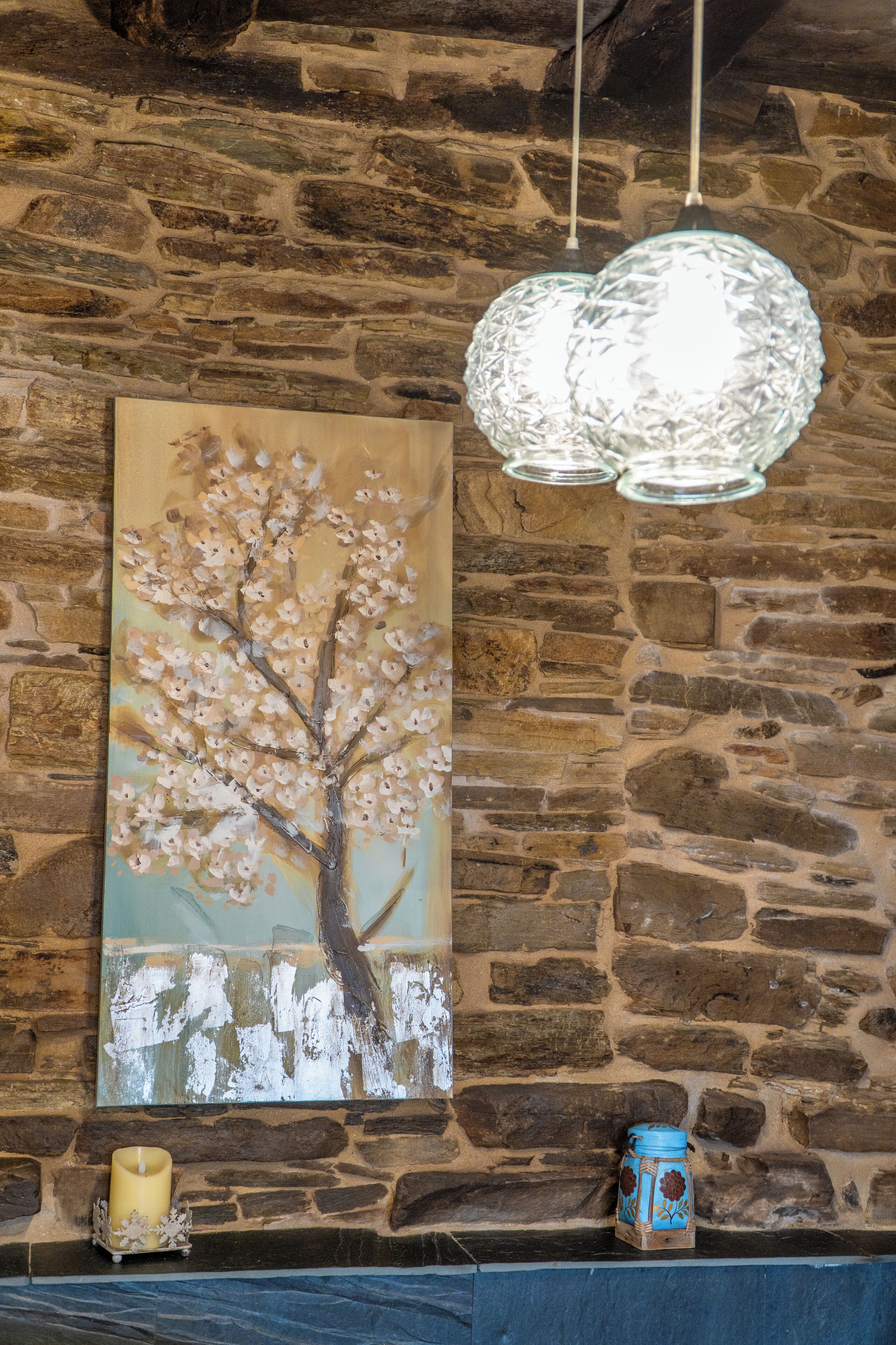 Pared de piedra con un cuadro de un árbol, una vela y un florero azul.