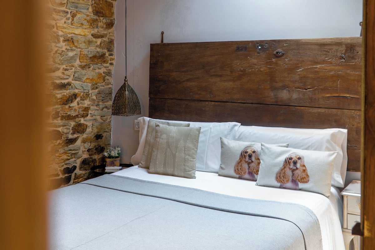 A bedroom with a sloped ceiling, exposed wooden beams, a stone wall, and a bed with white bedding and dog-themed pillows.