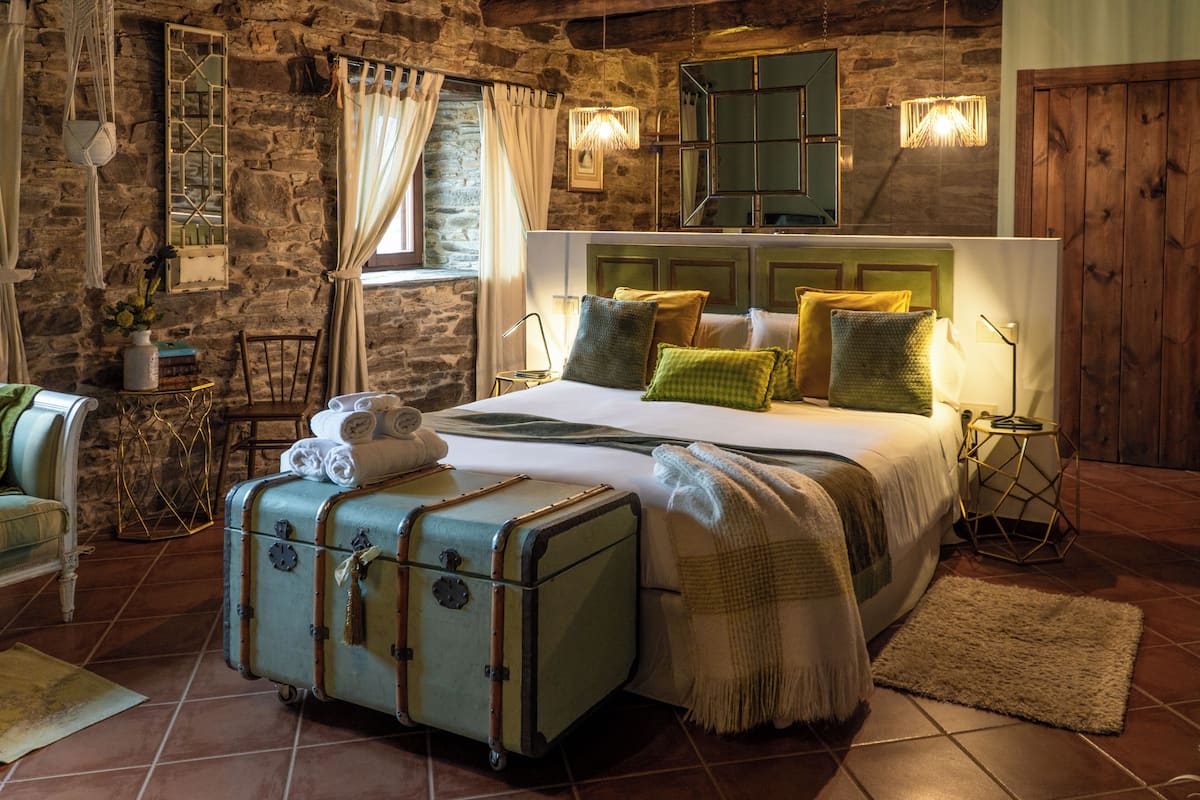 A rustic bedroom with a stone wall, a bed with multiple pillows, a suitcase, and a small table with books.