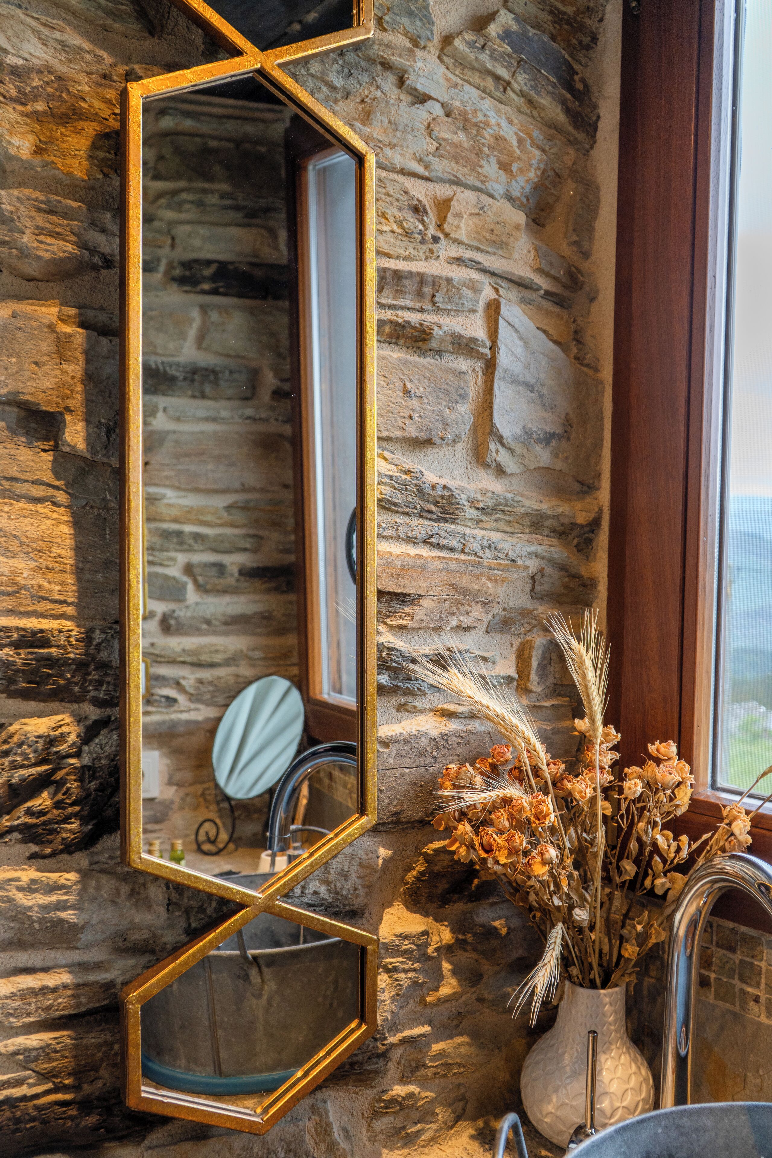 A bathroom with a stone wall, a gold-framed mirror, a sink, and a vase with dried flowers.