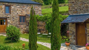A stone building with a stone pathway, two tall cypress trees, and a small garden area with a bench and chairs. - Complejo Rural Lar de Vies (A Pontenova)