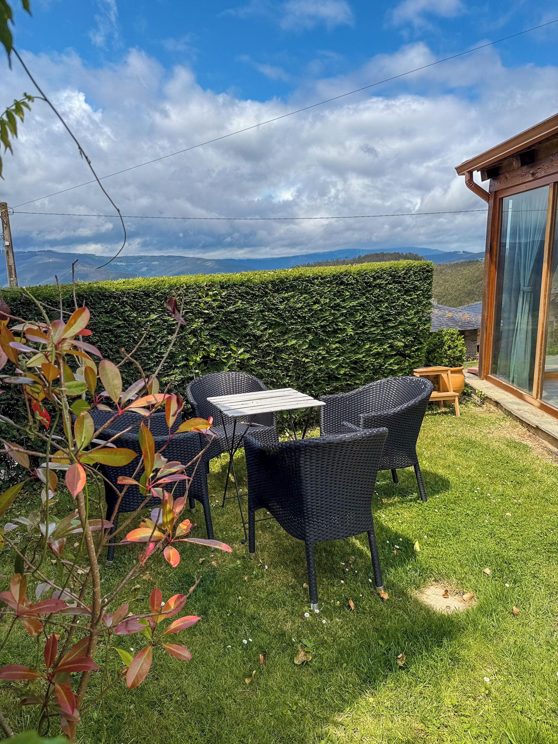 Een terras met rieten meubels en een tafel, met uitzicht op een keurig bijgesnoeid haagje en een verafgelegen heuvellandschap.