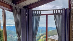 A cozy room with a grey sofa, a round table with a woven stool, and a view of a green landscape through large windows with curtains. - Complejo Rural Lar de Vies (A Pontenova)