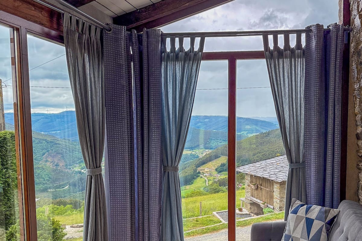 A cozy room with a grey sofa, a round table with a woven stool, and a view of a green landscape through large windows with curtains.