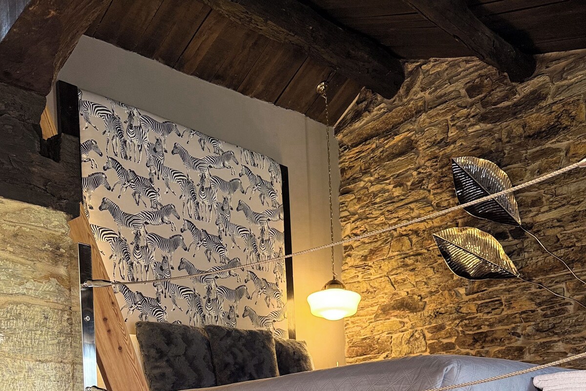 A loft-style bedroom with a wooden ceiling, stone walls, a zebra-patterned headboard, a hanging leaf light, and a bed with a grey quilt.