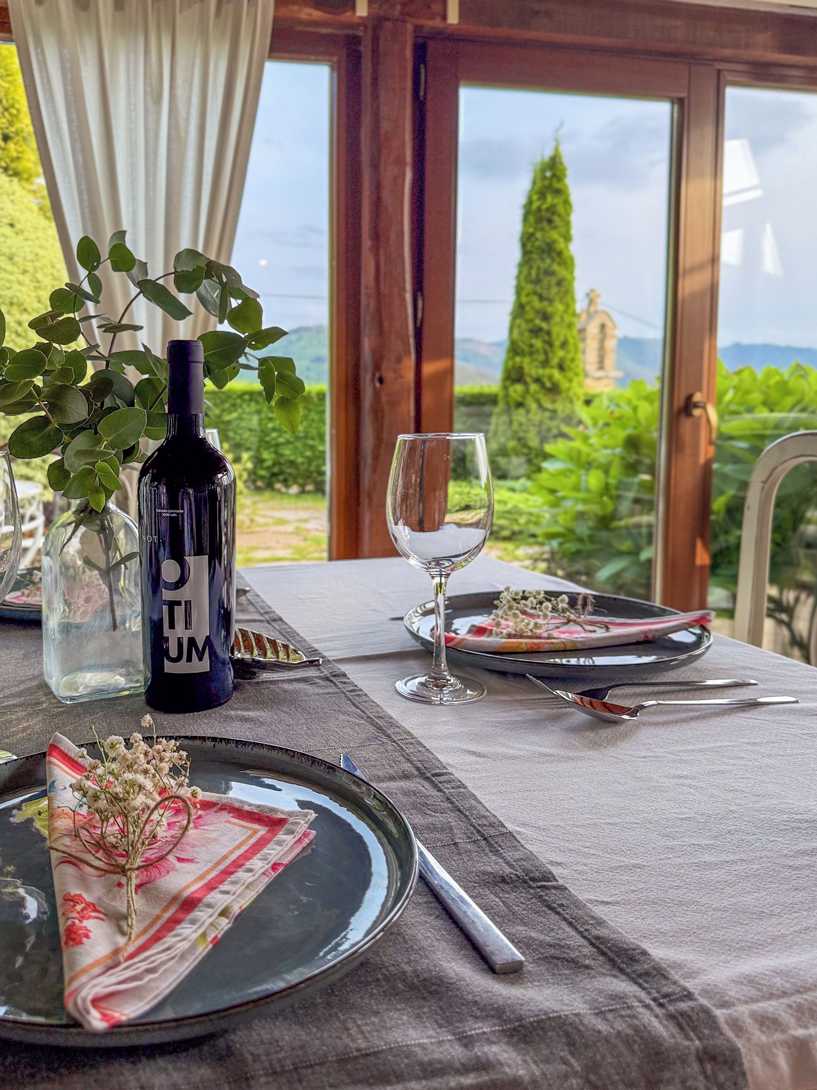 Una mesa dispuesta con una botella de vino, una copa, platos y cubiertos, con una vista panorámica a través de la ventana.