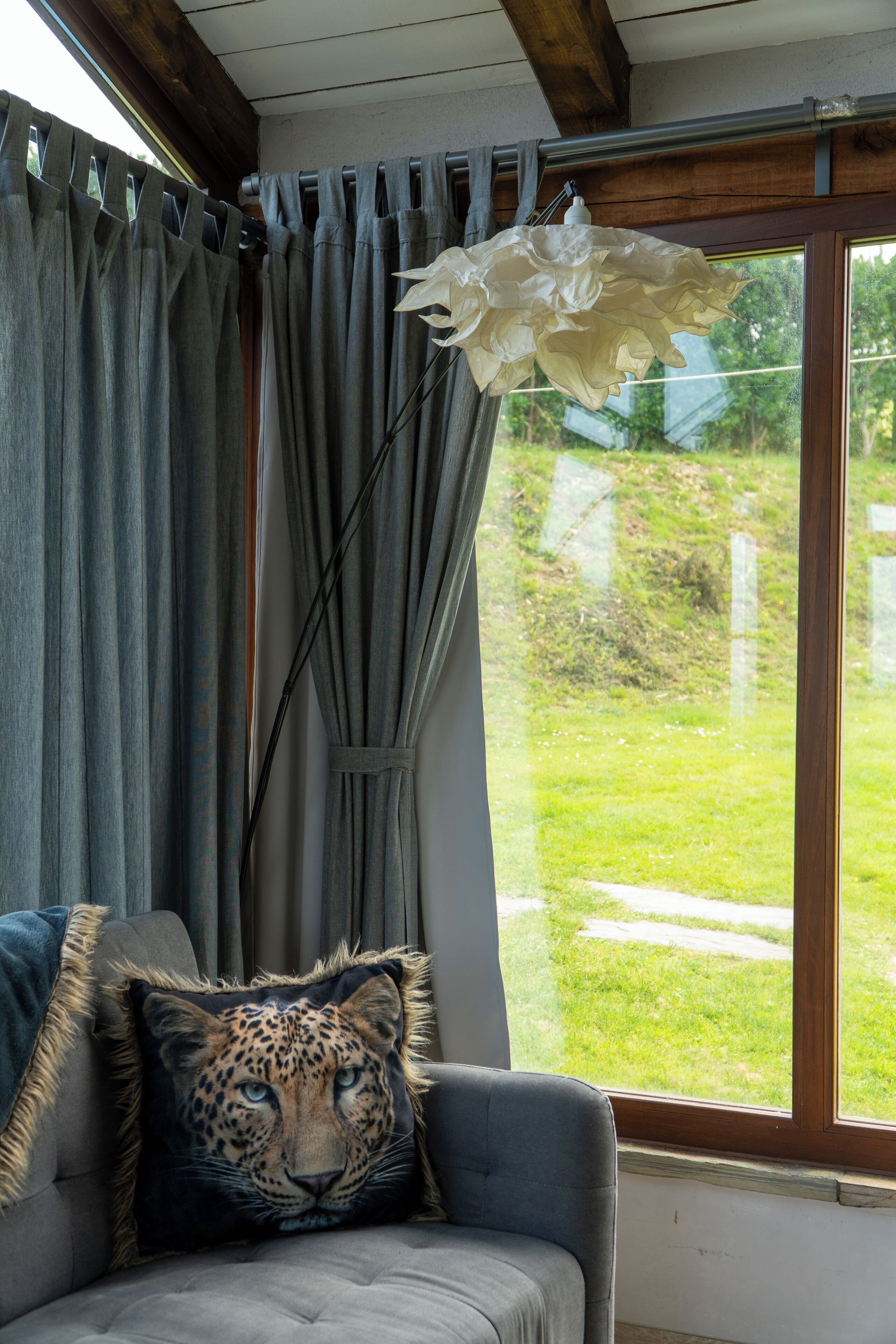 A room with a grey sofa, a leopard print pillow, grey curtains, and a window with a view of a green lawn.