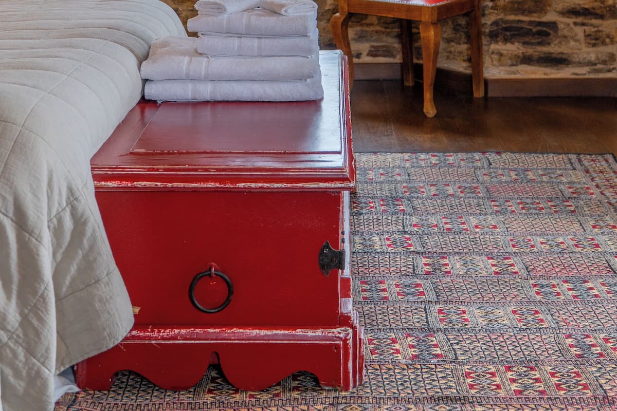 A room with a stone wall, a window with lace curtains, a wooden chair with a checkered cushion, a red chest of drawers, and a bed with a white duvet.