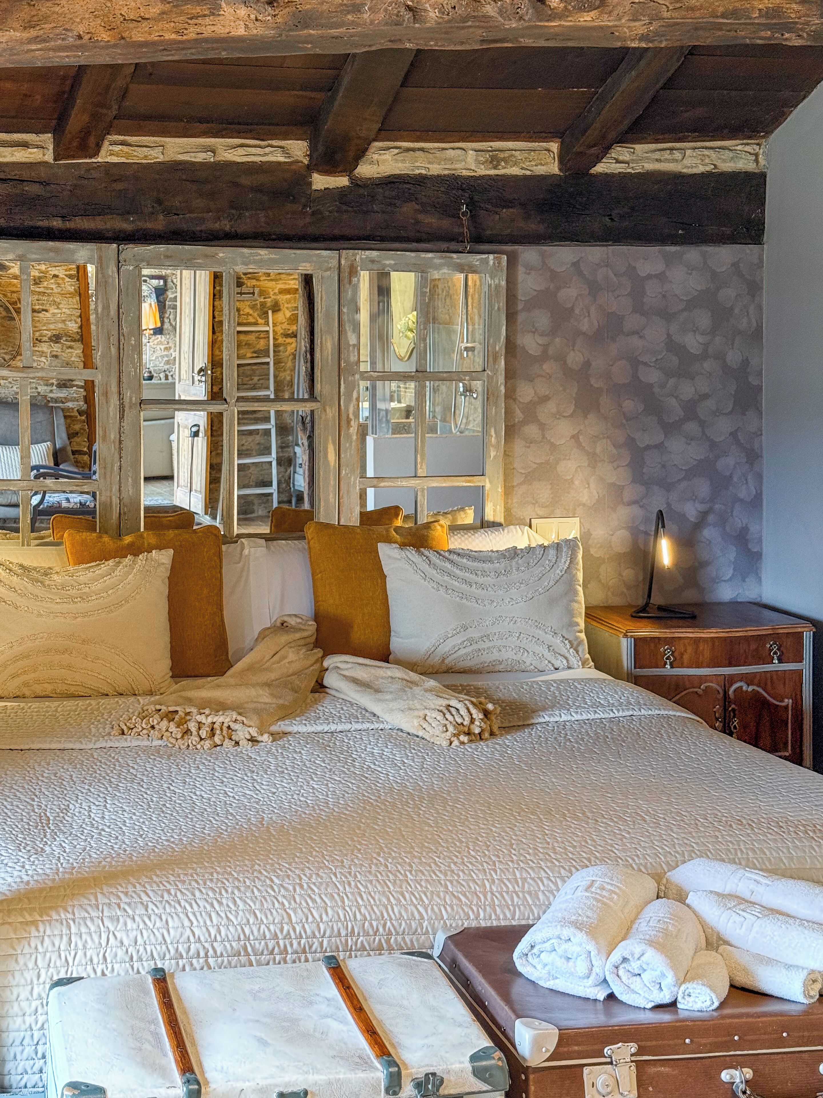A cozy bedroom with a large bed, wooden beams, stone walls, and a view through a window.
