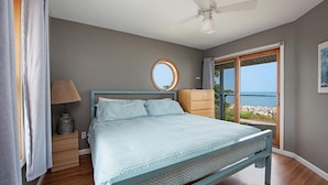 A bedroom with a bed, a nightstand, a dresser, a ceiling fan, a window with a view of the sea, and a circular mirror on the wall. - Lakefront 3-BR Home Summer Getaways & Sports Force (Sandusky)