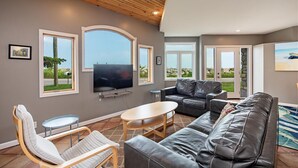 A modern living room with a large flat-screen TV, leather sofas, a coffee table, and large windows offering views of the outdoors. - Lakefront 3-BR Home Summer Getaways & Sports Force (Sandusky)