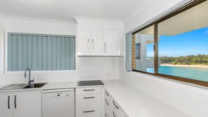 A compact kitchen with a built-in dishwasher, a window with blinds, and a view of a beach. - Creekfront Kingscliff Waters 2 (KINGSCLIFF)