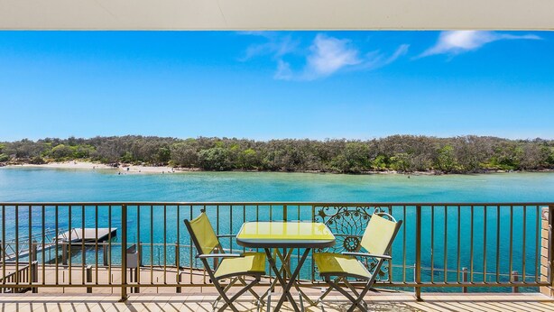 A balcony with a table and chairs overlooking a turquoise sea and a forested beach. - Creekfront Kingscliff Waters 2 (KINGSCLIFF)