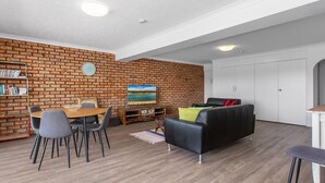 A living room with a brick wall, a dining area with a table and chairs, a black leather sofa, and a bookshelf. - Creekfront Kingscliff Waters 2 (KINGSCLIFF)