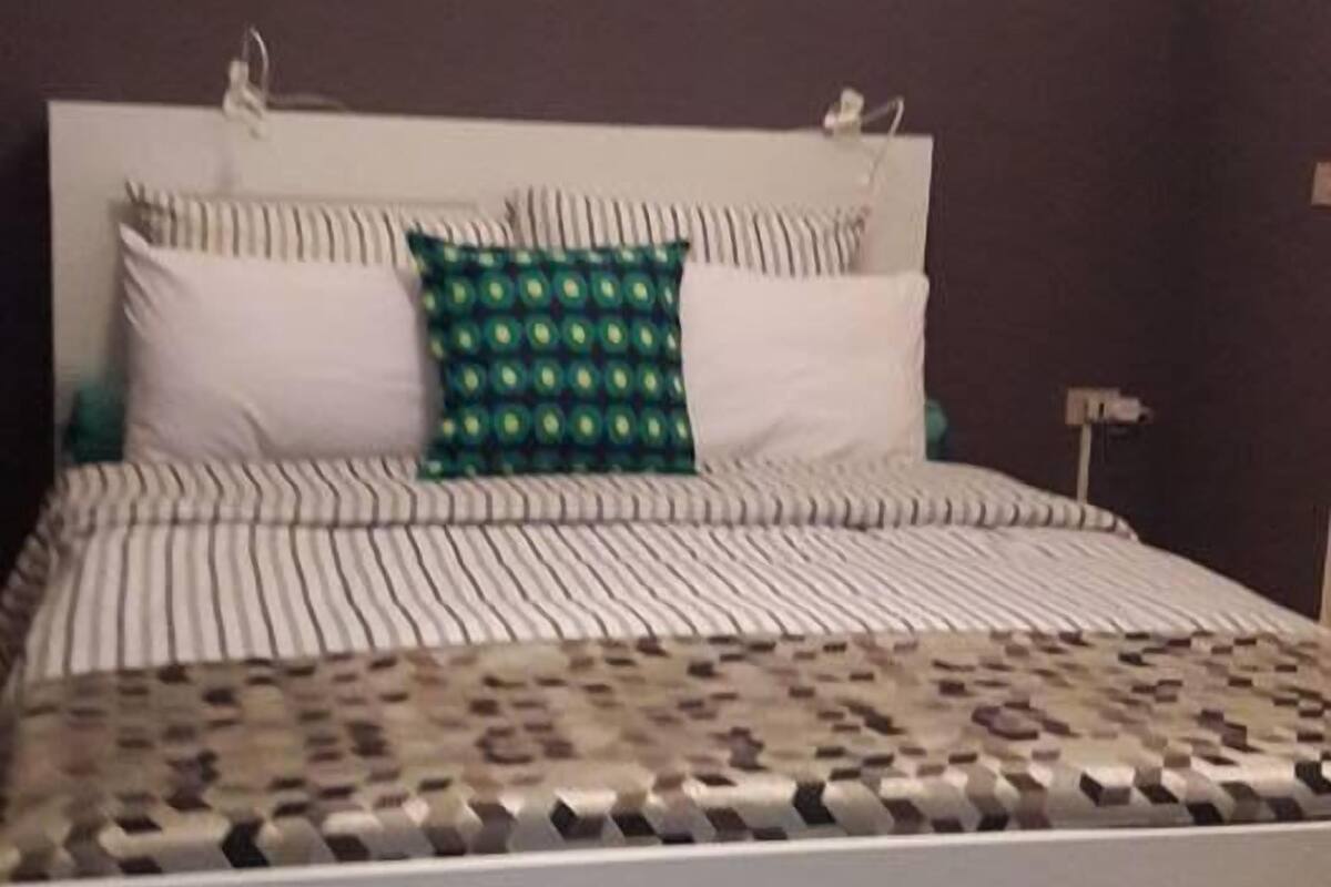 Ein Bett mit gestreifter Bettwäsche und einem grün gemusterten Kopfkissen, ein weißes Kopfteil und ein Metallgestell am Fußende des Bettes.