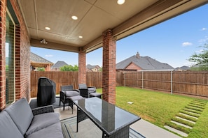 Une terrasse couverte, aménagée avec du mobilier d’extérieur, un barbecue et offrant une vue sur une cour arrière avec une clôture en bois et des maisons en arrière-plan.
