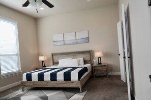A bedroom with a bed, bedside tables, a ceiling fan, a window with blinds, and a wall-mounted artwork.