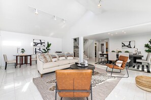 A spacious living area with a sofa, coffee table, and leather chairs. There is a dining area with a table and chairs in the background.