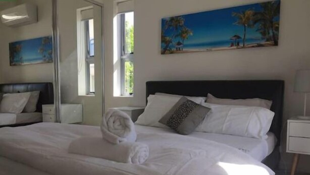 A neatly made bed with white linens and a rolled towel, a painting of a beach scene on the wall, and a window providing natural light. - Perch Apartments at Anembo Street (Surfers Paradise)