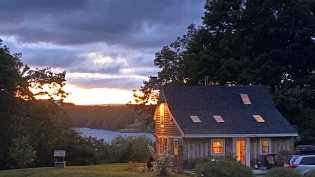 En rustik hytte med skråtag og ovenlys, omgivet af frodigt grønt og en sø.