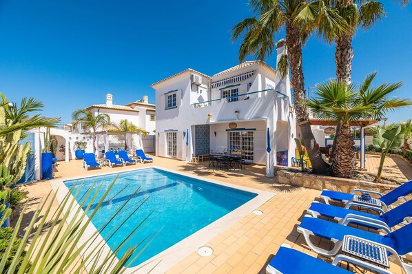 A poolside area with a clear blue pool, blue lounge chairs, and palm trees.