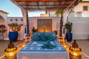 A bed with a patterned bedspread, surrounded by potted plants and lanterns, under a thatched roof structure.