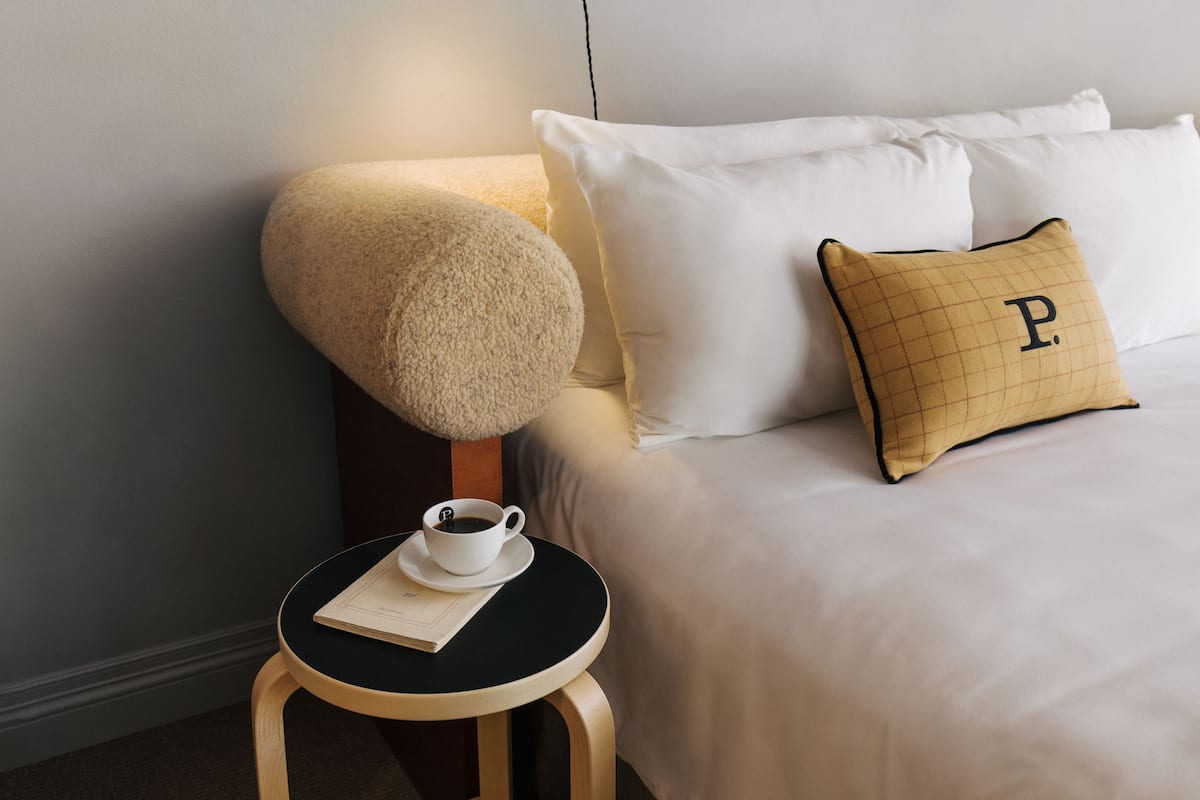 Una cama bien hecha con sábanas blancas y un almohadón de estampado en cuadrícula con una 'P' grande. Una mesita redonda con tapa negra sostiene una taza y un platillo blancos, junto con un menú o folleto.