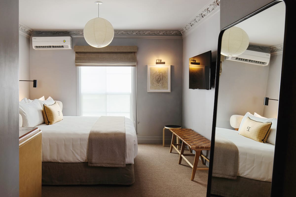 Una habitación de hotel con una cama grande, una mesita de noche, un banco, un televisor montado en la pared y un equipo de aire acondicionado.