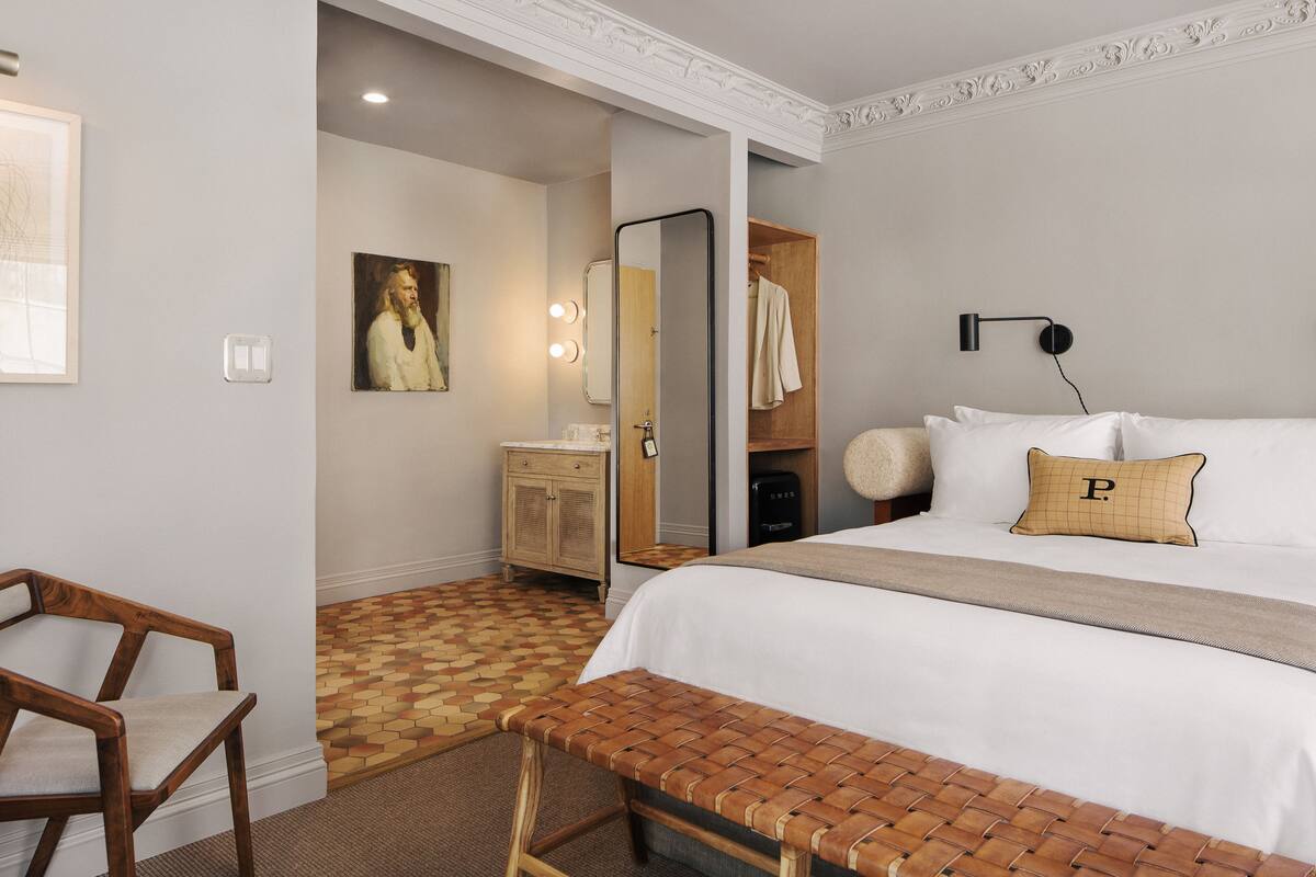 Habitación de hotel con cama, banca, aparador de madera, espejo y lámpara de pared.