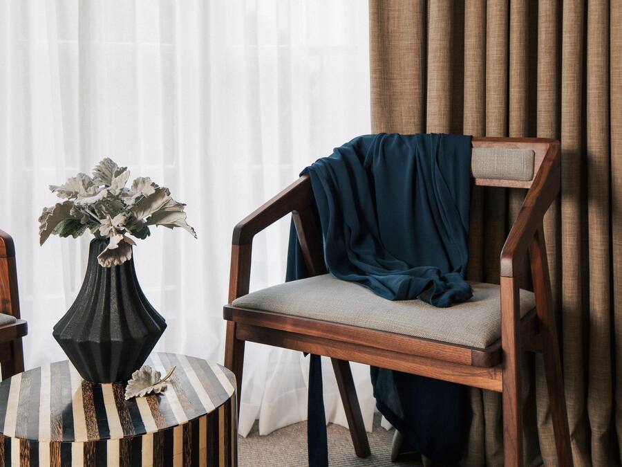 Een kamer met een houten stoel, een rond bijzettafeltje met strepen, een vaas met bloemen, en een paar schoenen op de vloer.