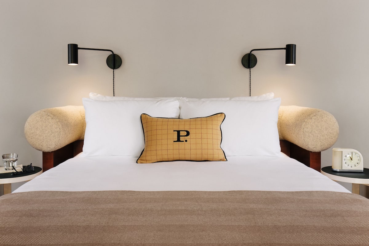 Una cama bien hecha con ropa de cama blanca y marrón, un cojín con la letra 'P', dos lámparas de noche redondas y una mesita de noche con un reloj y un vaso de agua.