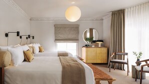 Ein Hotelzimmer mit zwei Betten, einer Holzbank, einem Schreibtisch, einem Stuhl und einem großen Fenster mit Vorhängen.