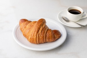 Een croissant op een wit bord, met een kop koffie en een lepel op een schoteltje.