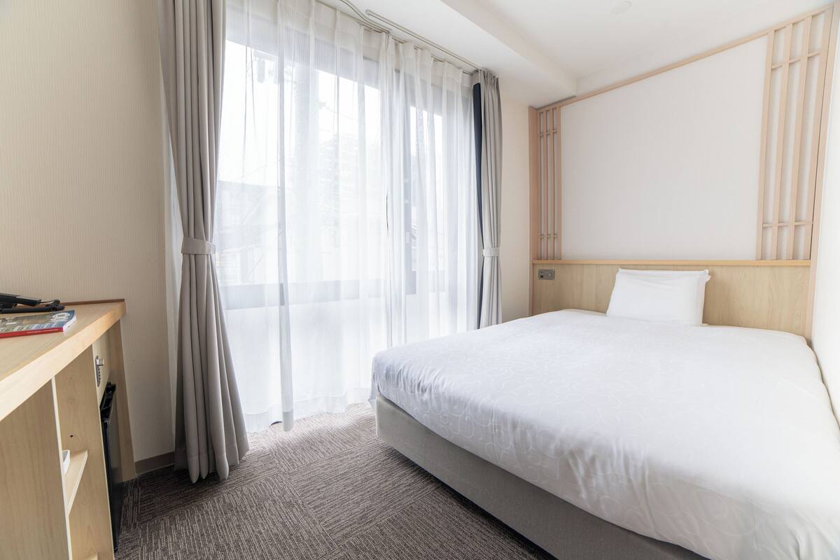 Et hotelværelse med seng, skrivebord og et vindue med let gardin.