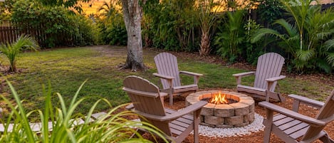 A fire pit with four Adirondack chairs arranged around it, surrounded by a grassy area with trees and plants.