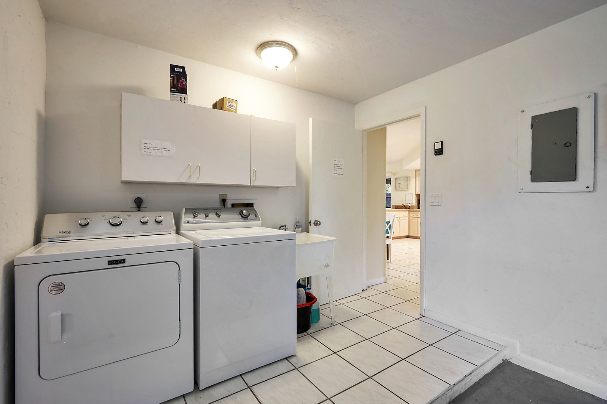 A laundry room with two front-loading washing machines and a dryer, a white cabinet above, and a tiled floor.