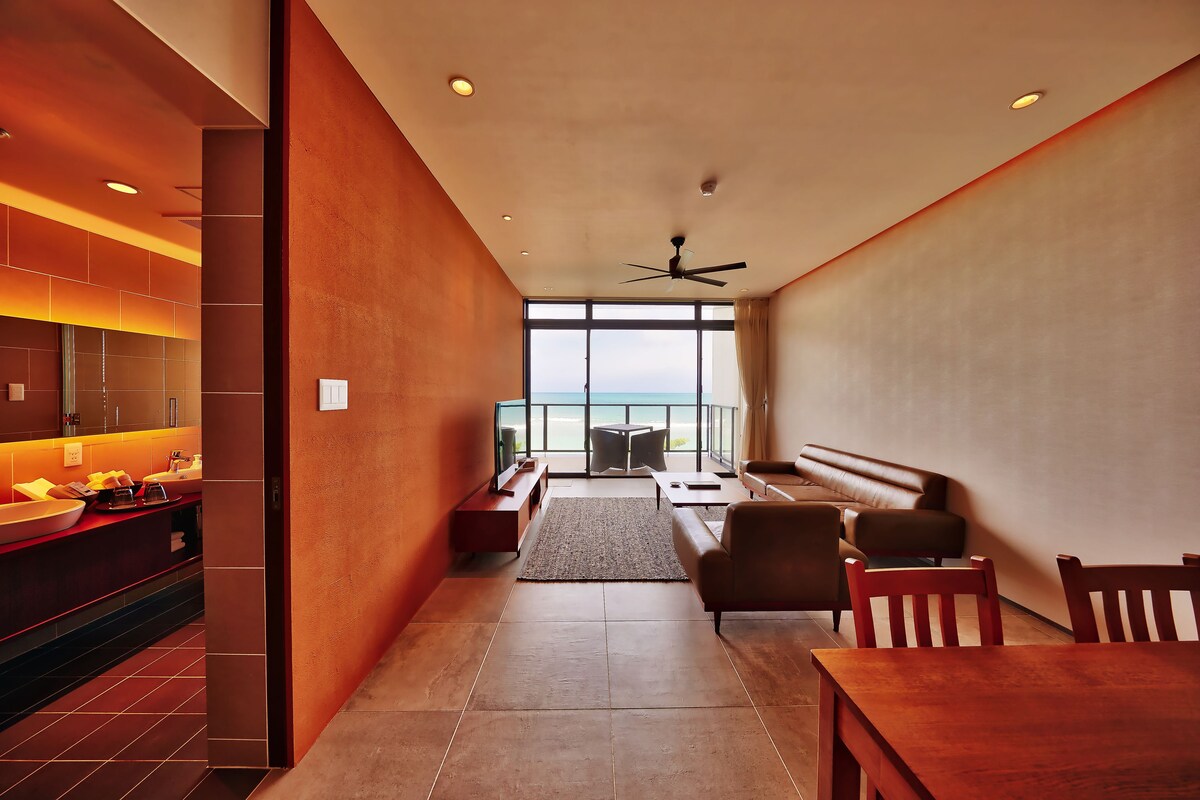 A modern hotel room with a kitchen area, a seating area with a sofa and chairs, and a large window offering a view of the sea.