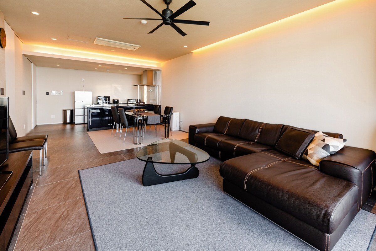 A modern living room with a leather sofa, a glass coffee table, and a dining area in the background.