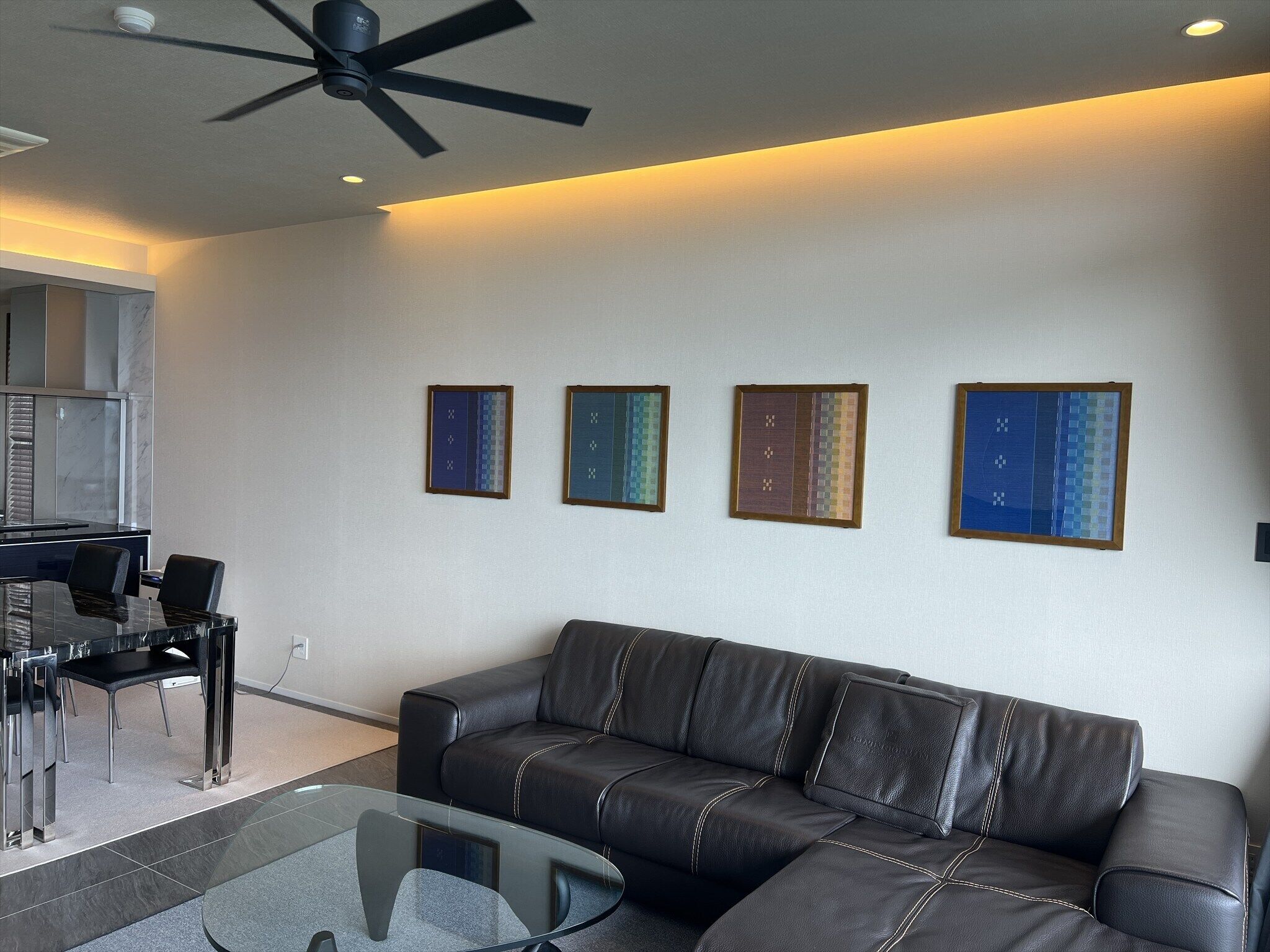 A modern living room with a leather sofa, a glass coffee table, and a ceiling fan. There are four framed artworks on the wall.