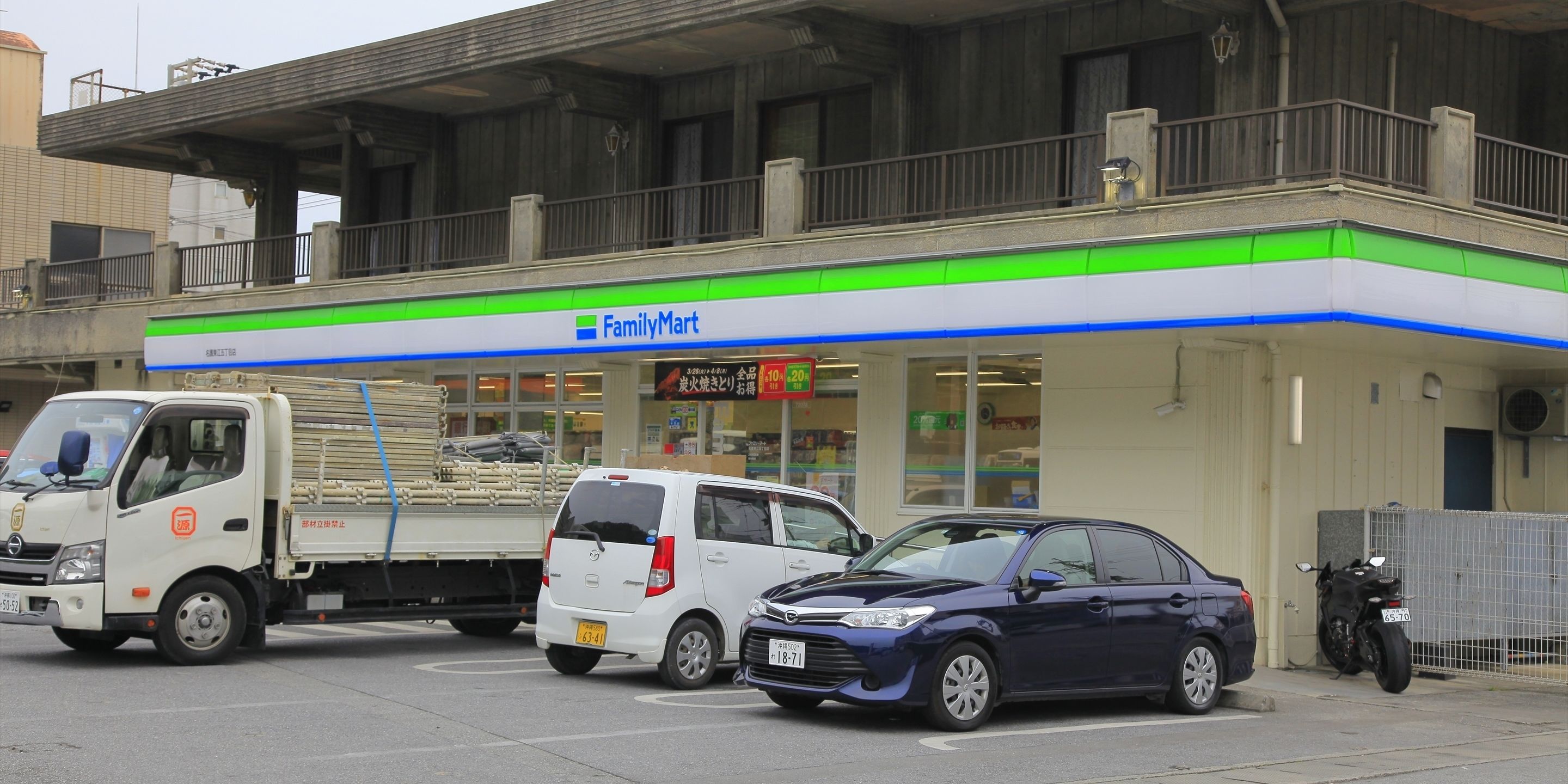 Una tienda FamilyMart con fachada blanca y verde, un estacionamiento con varios vehículos y una motocicleta estacionada afuera.