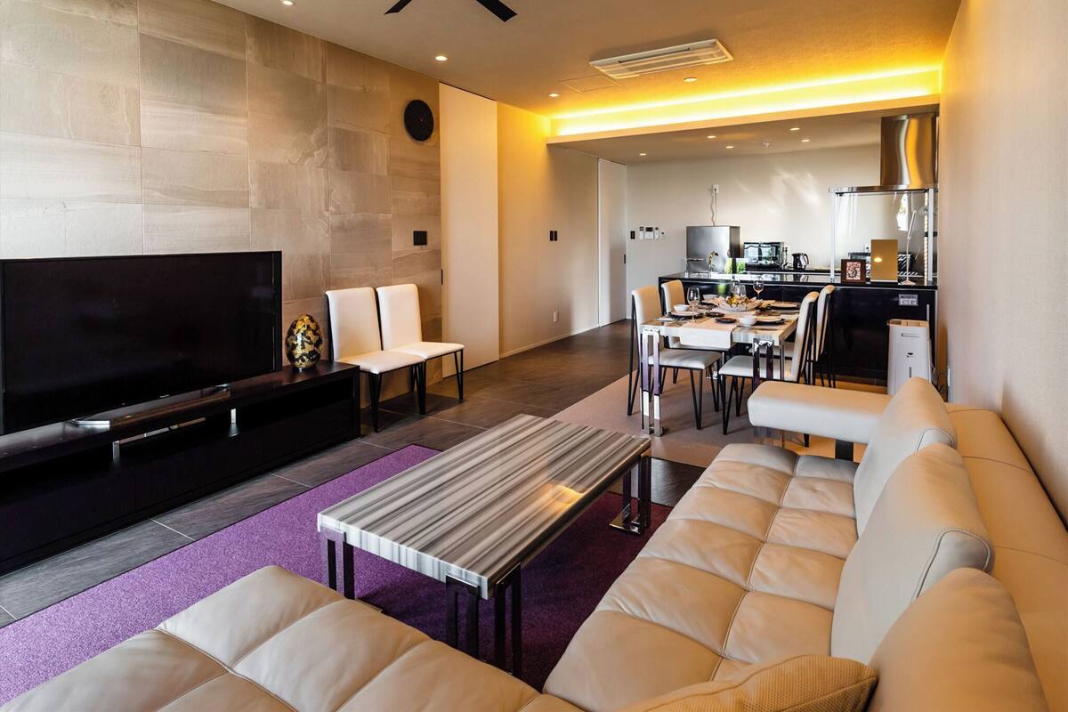 A modern living room with a flat-screen TV, a ceiling fan, a dining area with a kitchen in the background, and a purple rug.