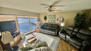 A living room with a large TV, leather sofas, a ceiling fan, and a view of a body of water through large windows.